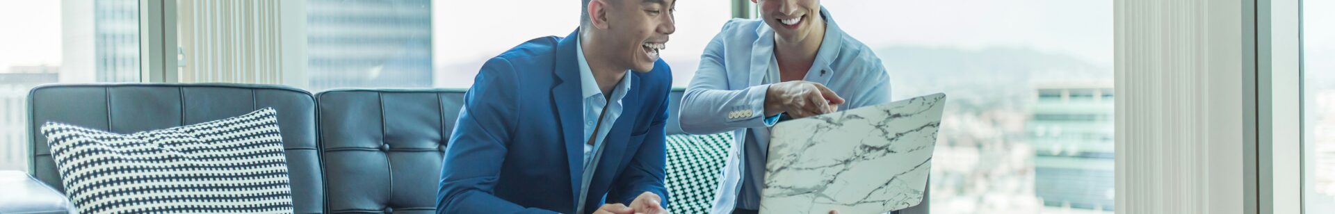 two men sitting on a sofa in an office and cheerfully looking at a laptop screen