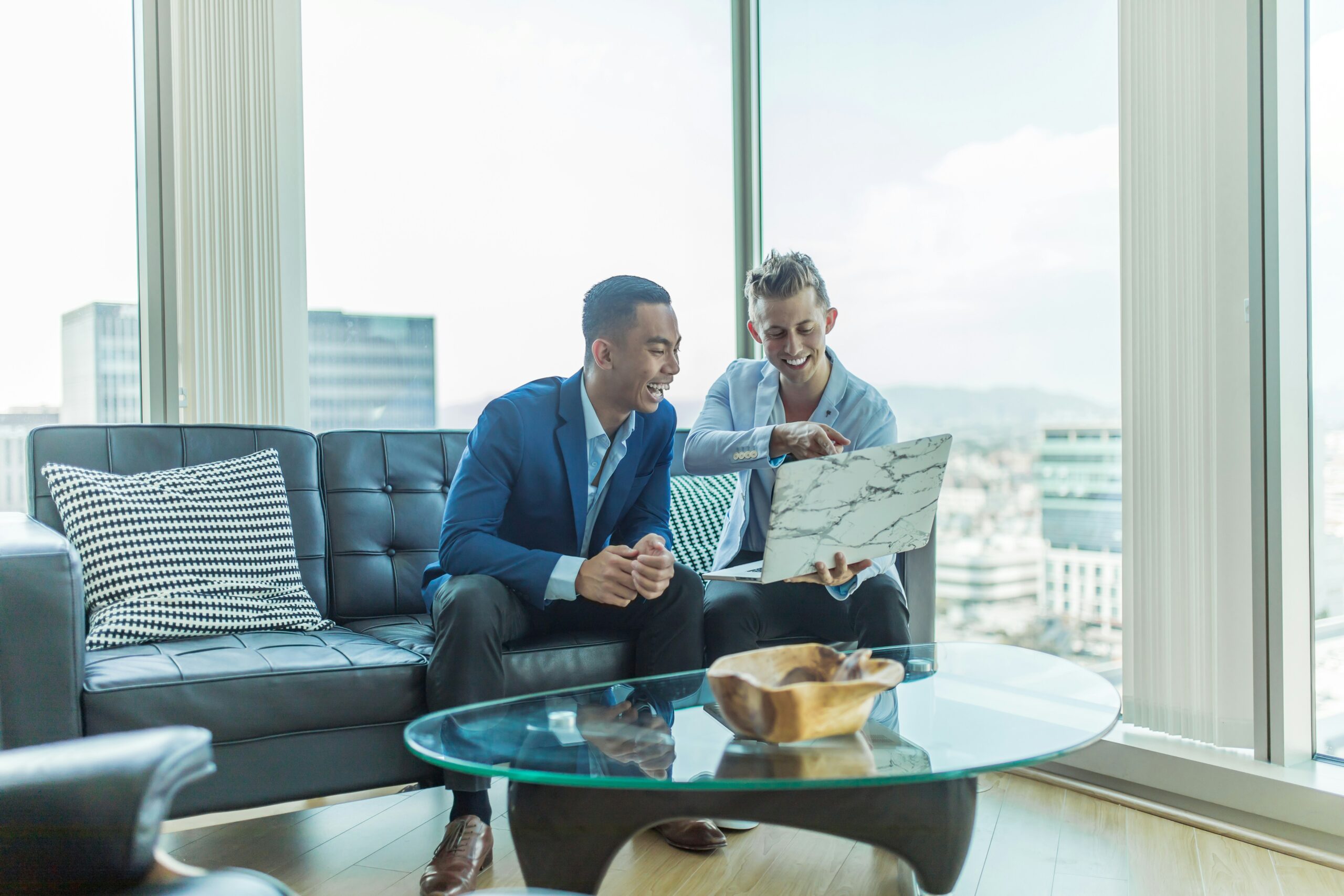 two men sitting on a sofa in an office and cheerfully looking at a laptop screen