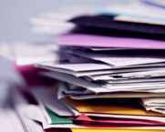 A tall, disorganised stack of mixed paper business documents and coloured folders in close-up, representing the manual document handling that cbs AID and SAP Business AI convert into SAP-ready data.
